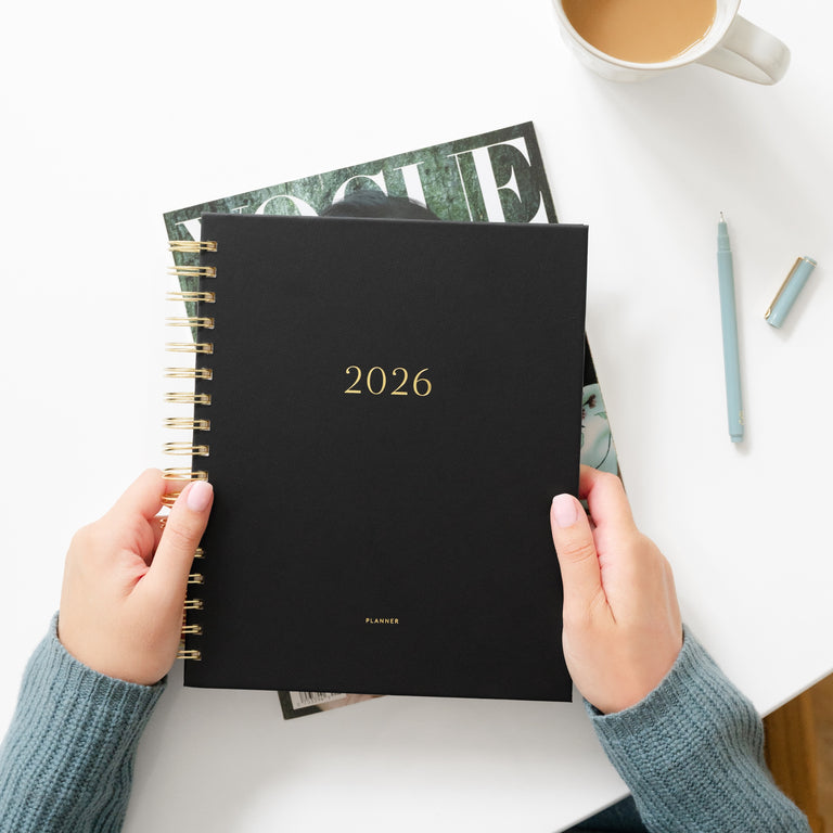Person holding a black planner with '2026' on it, placed on a white surface with a cup of coffee