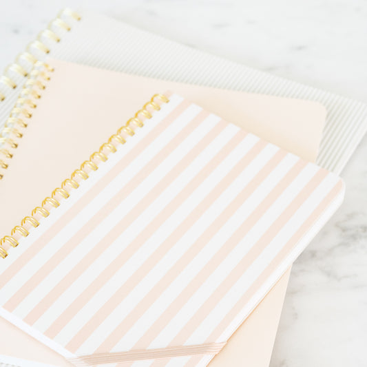 A stack of spiral bound notebooks in three sizes in pink, white and grey colors with gold spiral binding