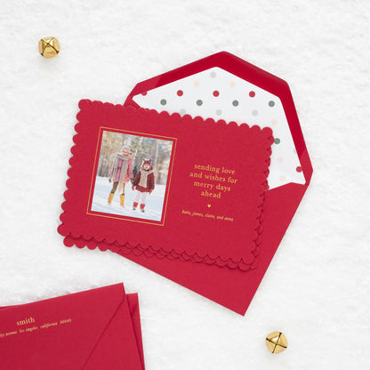 Close up of red scalloped custom holiday photo card with a photo of two kids in the snow with gold foil 'sending love and wishes for merry days ahead' paired with stack of red envelopes