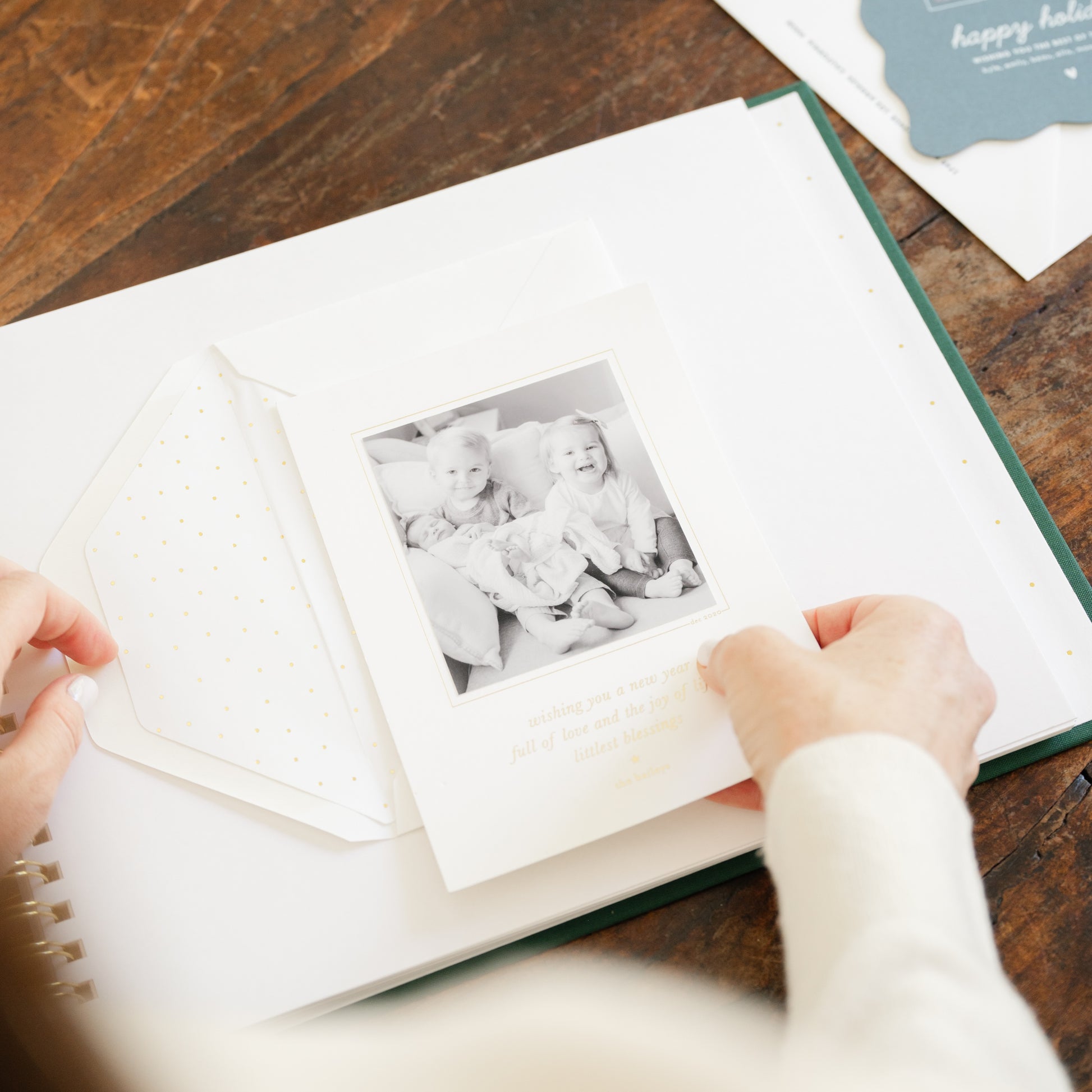 A person's hands holding a holiday card with a black-and-white photo of two children, placed over an open white album or envelope on a wooden table.