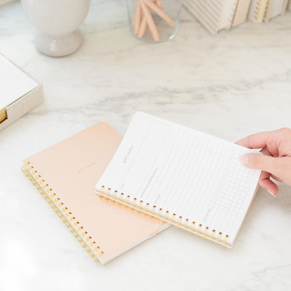 Pink Goal Tracker Spiral Notebook open with guided prompts in hand with a closed notebook in background