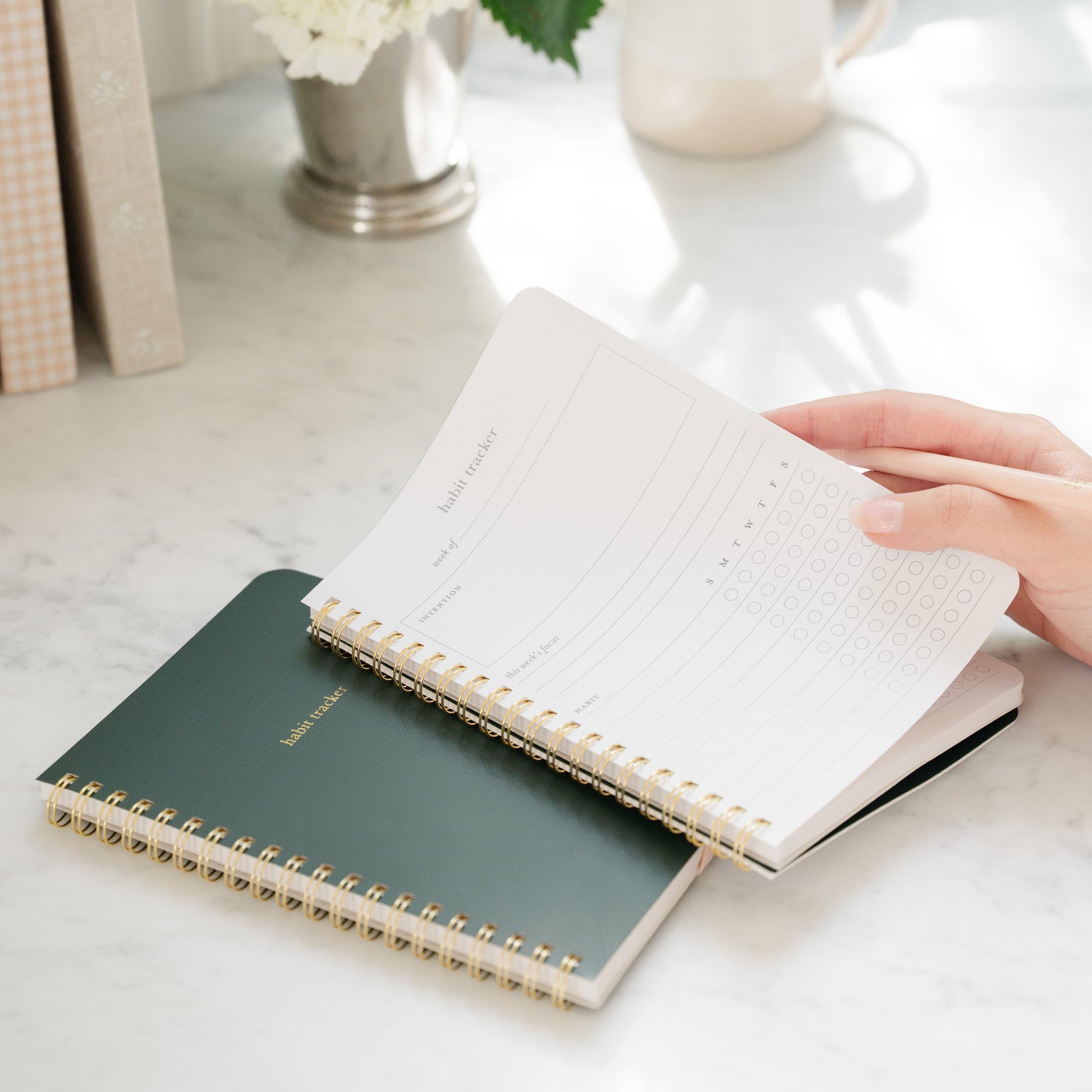 Green habit tracker spiral notebook closed in background with an open habit tracker notebook in hand with guided prompts on a kitchen counter
