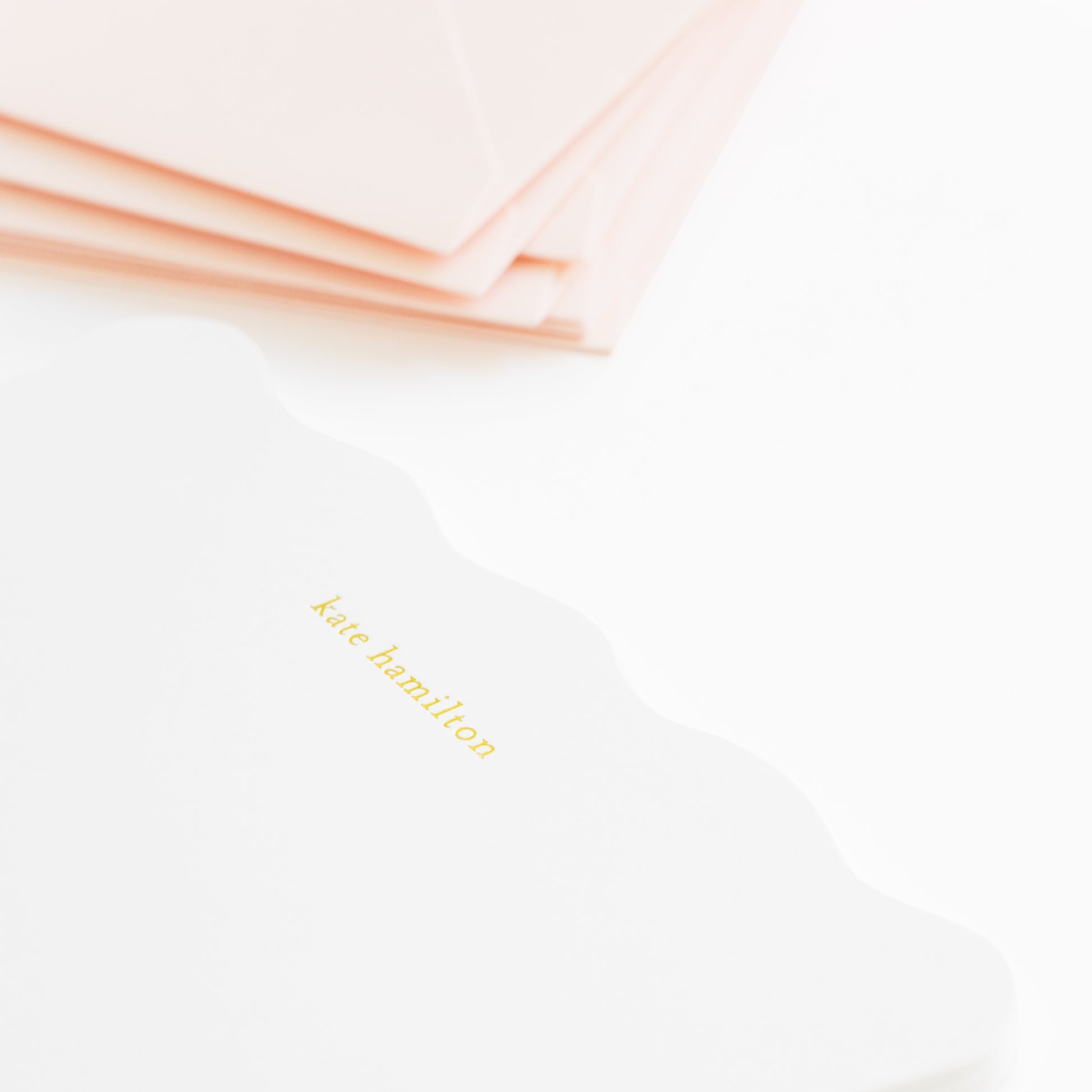 Stack of pale pink envelopes with white scalloped flat cards with 'kate hamilton' printed in gold foil