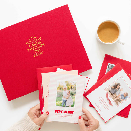 Red book labeled 'Our Holiday Cards Through the Years' with Christmas cards and a cup of coffee on a white surface.