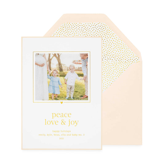 White custom holiday photo card printed with "peace love and joy" in gold foil with a photo of two kids and parents with a pink card border paired with a pink envelope open to gold teensy dot envelope liner